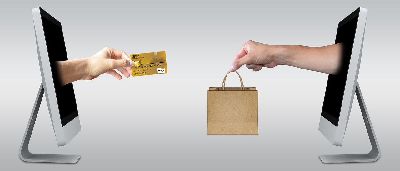 Two hands reaching through computer screens, one handing over a credit card and the other receiving a paper shopping bag filled with CreatingPrintables.