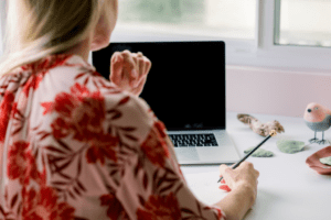 A woman in a floral blouse works at a laptop, holding a pencil and making notes on compatibility warnings, with decorative items on the desk.