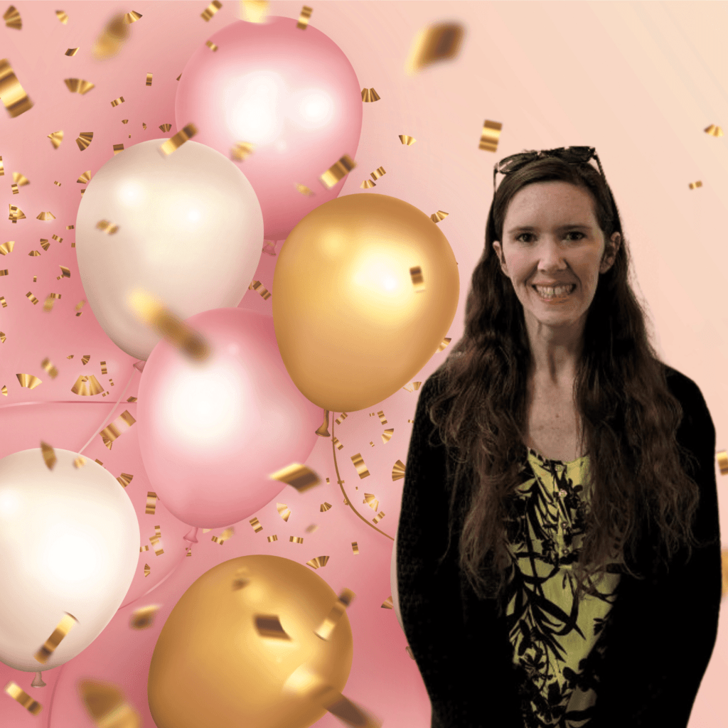 A woman with long hair stands smiling in front of a backdrop featuring pink, white, and gold balloons with golden confetti. She is wearing a yellow and black patterned top with a black cardigan.