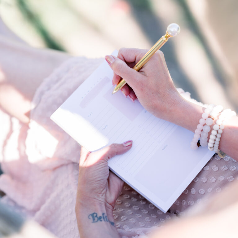 A person practicing self care journaling wears a pink dress and beaded bracelets, writing in a planner with a gold pen while sitting outdoors.