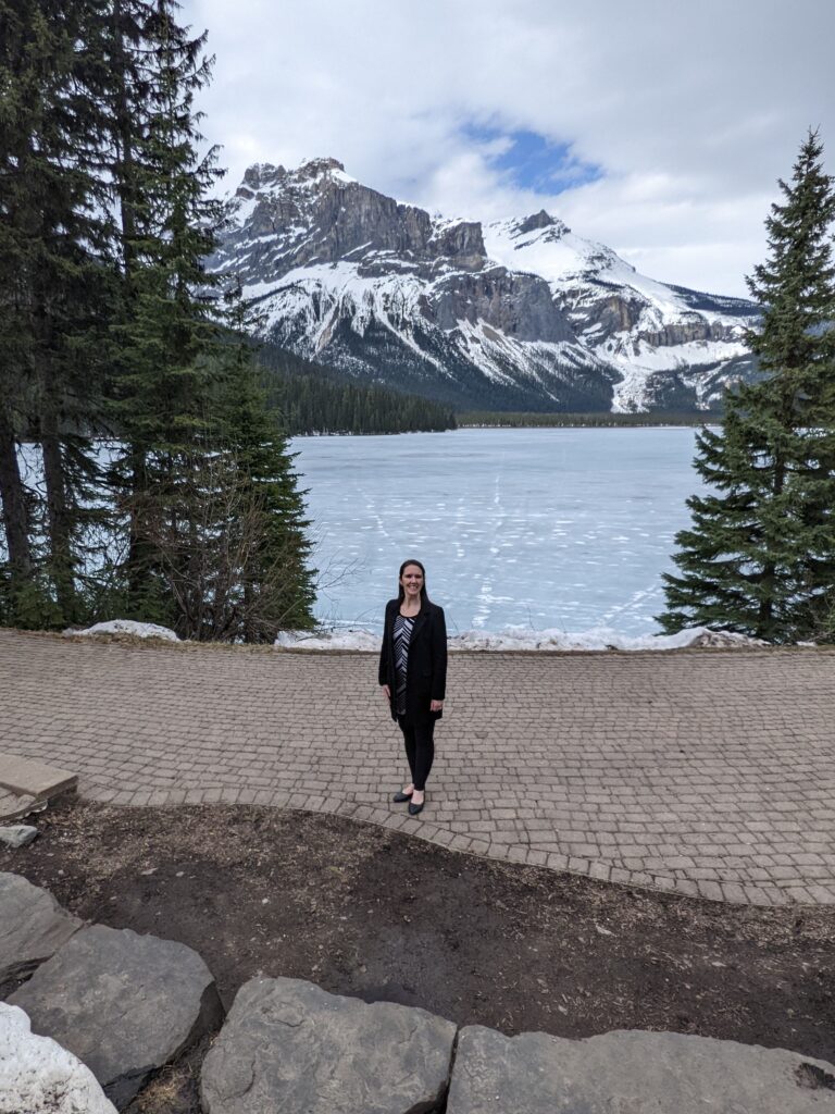 A business mentor for creatives stands on a paved path in front of a partially frozen lake, with evergreen trees and snow-capped mountains in the background—an inspiring scene for any Business Mentor for Creatives seeking fresh ideas.