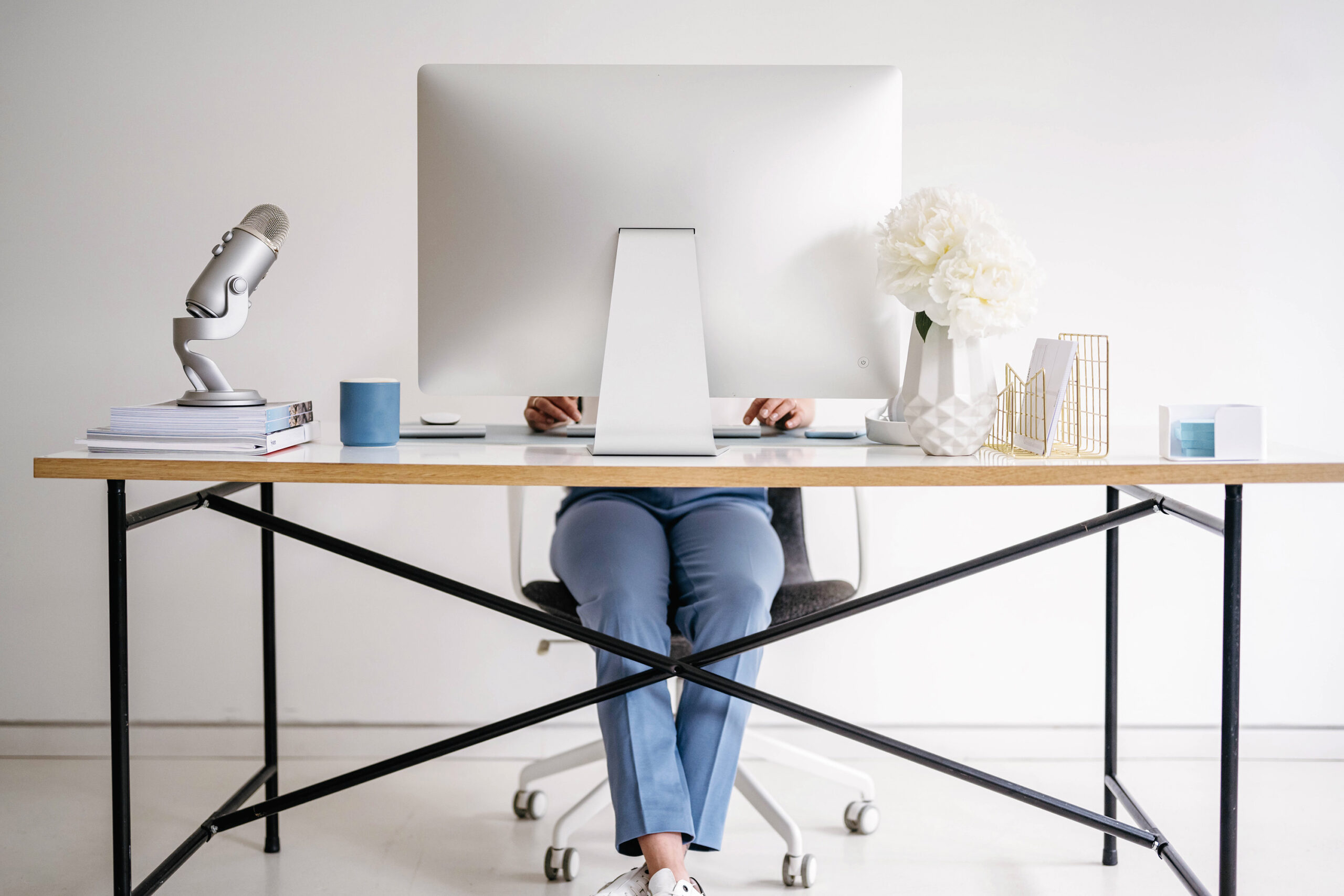 A Business Mentor for Creatives sits at a desk using a computer to create PLR & MRR templates, with a microphone, books, cup, and decorative flowers arranged on the desktop against a white background.