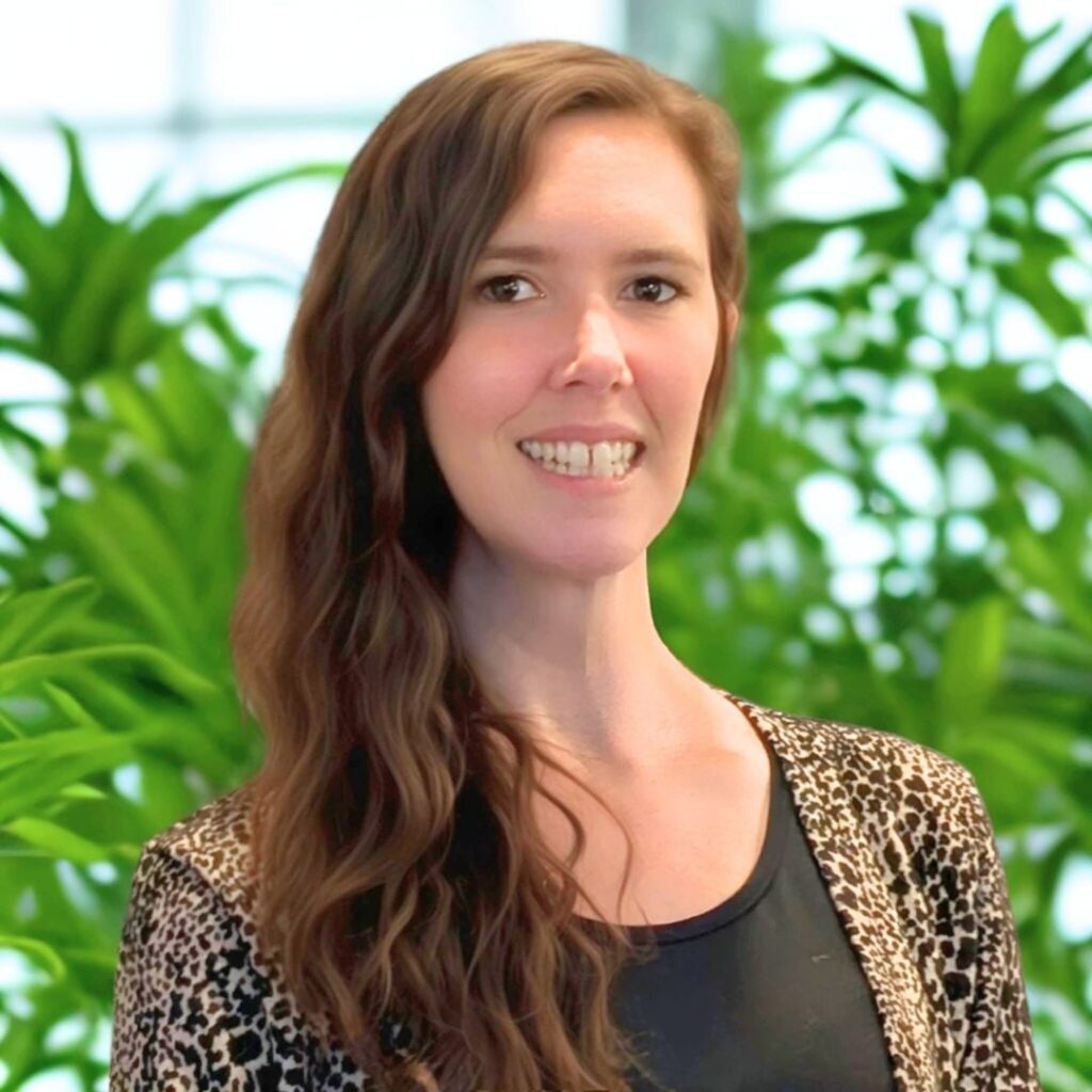 A picture of Aimee with long, wavy brown hair and a leopard print cardigan stands in front of green leafy plants, smiling at the camera